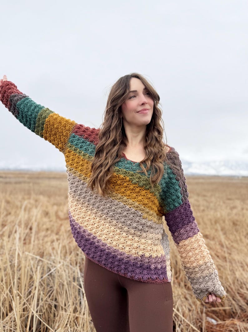 May include: A woman wearing a multi-coloured crochet sweater with a diagonal stripe pattern. The sweater is made with a textured stitch and features shades of brown, green, purple, and yellow.