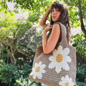 May include: A brown crocheted tote bag with white daisy flowers. The flowers have yellow centres.