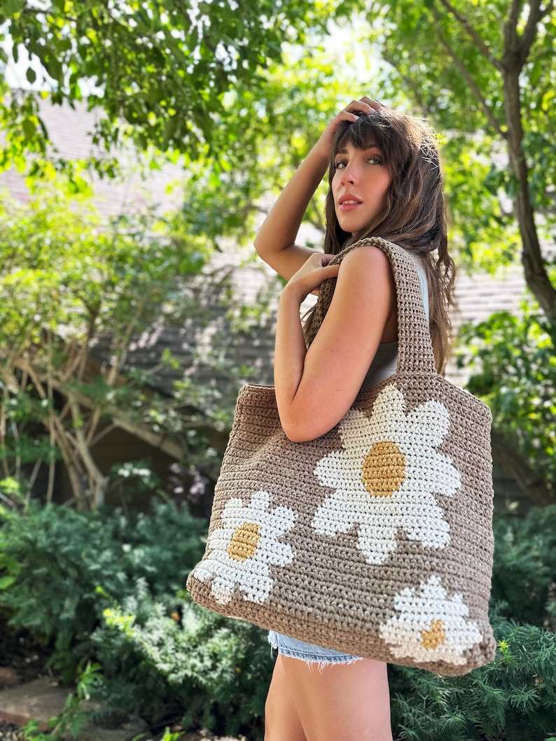 May include: A brown crocheted tote bag with white daisy flowers. The flowers have yellow centres.