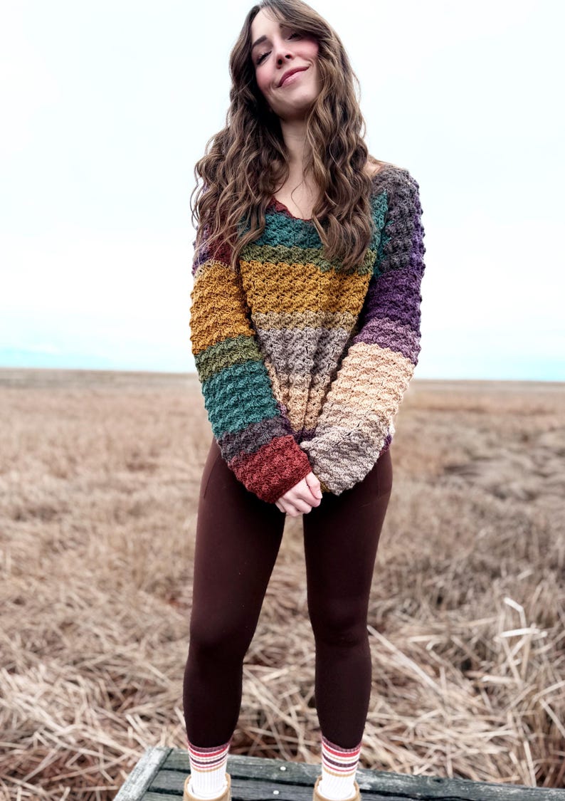 May include: A woman wearing a multi-coloured striped crochet sweater with brown leggings and white socks with colourful stripes.