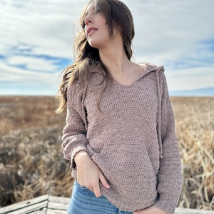 May include: A brown crocheted hooded sweater with a relaxed fit. The sweater has a V-neck and a drawstring hood.
