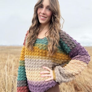 May include: A multi-coloured crochet sweater with a striped pattern in shades of brown, green, yellow, beige, and purple. The sweater has a relaxed fit and long sleeves.
