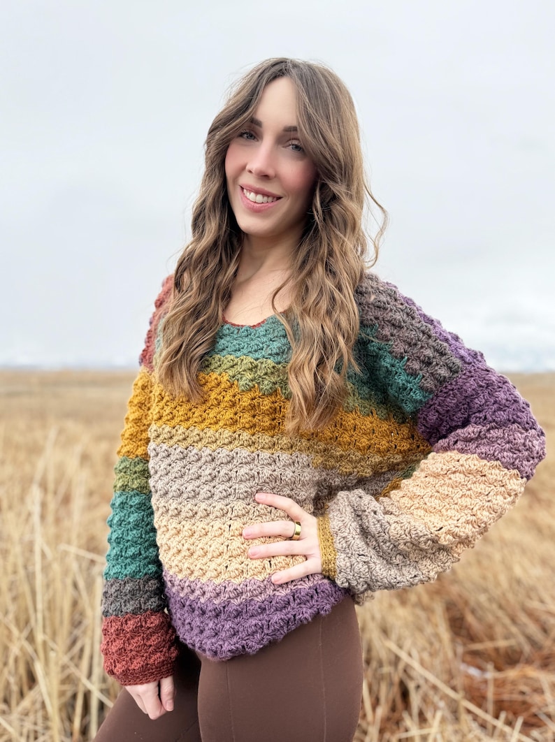May include: A multi-coloured crochet sweater with a striped pattern in shades of brown, green, yellow, beige, and purple. The sweater has a relaxed fit and long sleeves.
