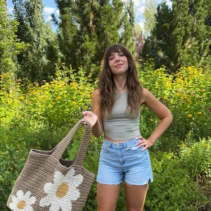May include: A brown crocheted tote bag with white daisy flowers. The bag has two handles and is large enough to carry groceries or other items.