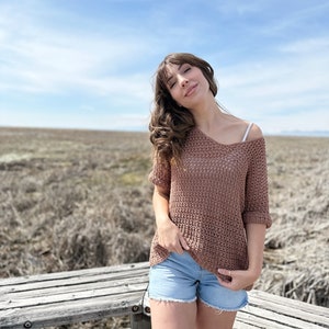 Puede incluir: Una mujer con un top de crochet marr&oacute;n y pantalones cortos de mezclilla azul se para frente a un banco de madera. El fondo es un campo de hierba seca y un cielo azul.