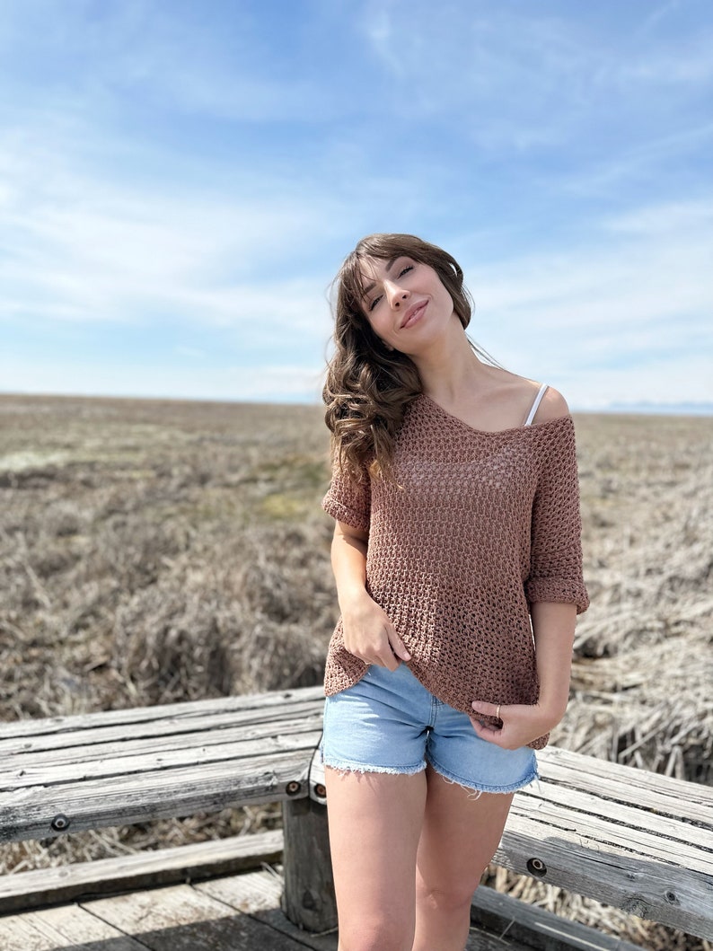 Puede incluir: Una mujer con un top de crochet marr&oacute;n y pantalones cortos de mezclilla azul se para frente a un banco de madera. El fondo es un campo de hierba seca y un cielo azul.