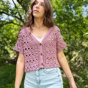 May include: A short-sleeved, button-down crochet cardigan in a dusty rose color. The open weave design reveals a white undershirt. The cardigan has a V-neck and is paired with light blue denim shorts. The background is a lush green.