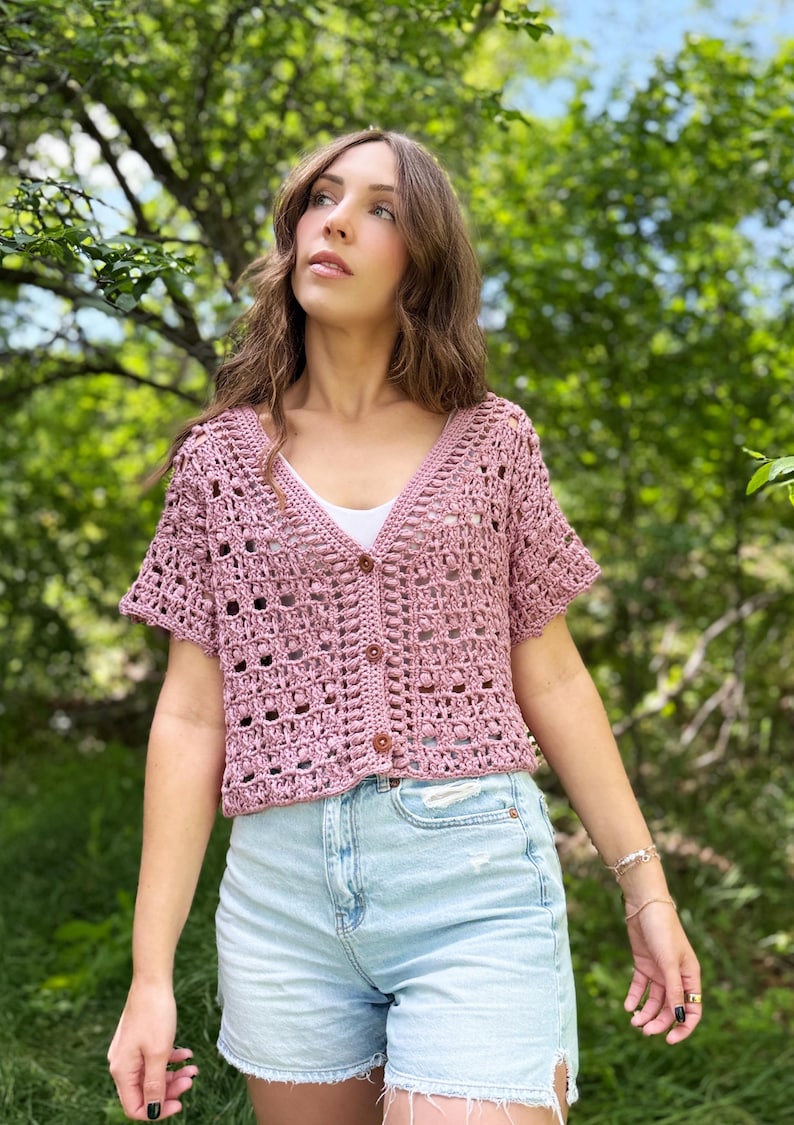 May include: A short-sleeved, button-down crochet cardigan in a dusty rose color. The open weave design reveals a white undershirt. The cardigan has a V-neck and is paired with light blue denim shorts. The background is a lush green.