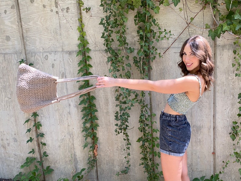 Puede incluir: Una mujer con un top corto de flores verdes y pantalones cortos de mezclilla sostiene una bolsa de mano de crochet beige.