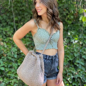 Puede incluir: Una mujer lleva un top corto de flores verdes y pantalones cortos de mezclilla y lleva un bolso grande de crochet beige con cierre de cord&oacute;n.