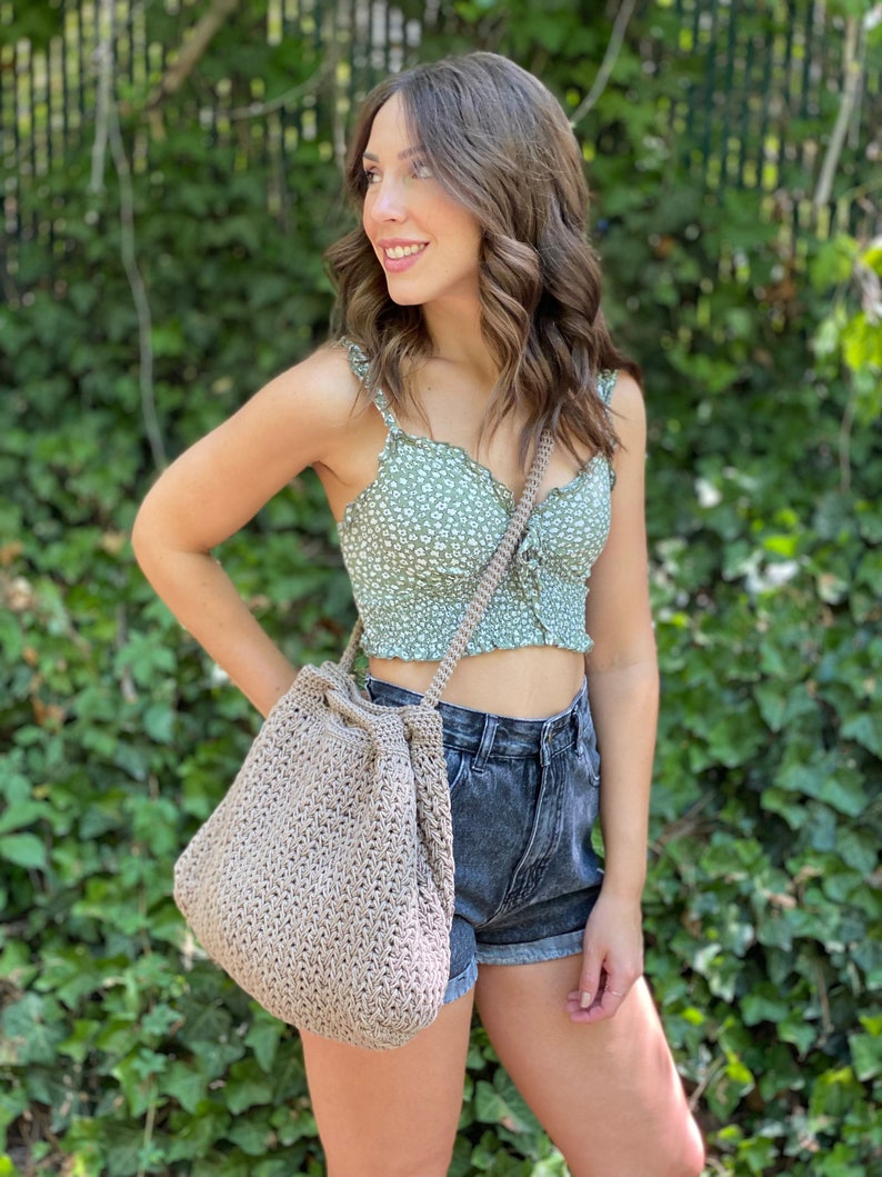 Puede incluir: Una mujer lleva un top corto de flores verdes y pantalones cortos de mezclilla y lleva un bolso grande de crochet beige con cierre de cord&oacute;n.