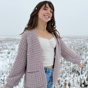 May include: A woman wearing a light purple crocheted cardigan with a white tank top and blue jeans. She is standing in a snowy field with her arms outstretched.