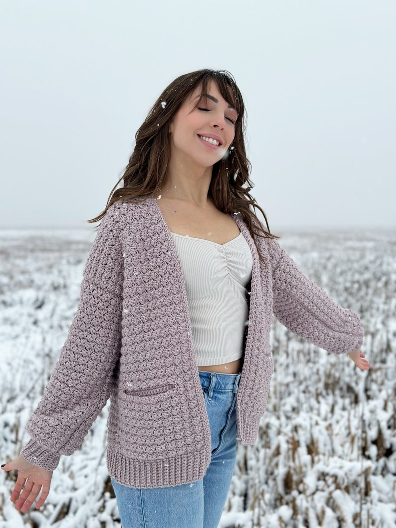May include: A woman wearing a light purple crocheted cardigan with a white tank top and blue jeans. She is standing in a snowy field with her arms outstretched.