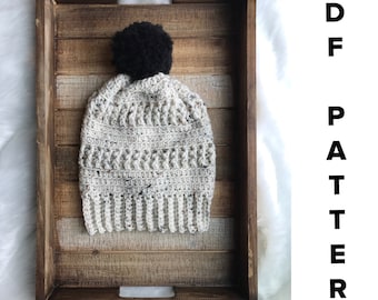 Puede incluir: Un gorro de punto de ganchillo con un pompón negro. El gorro es blanco con un patrón moteado y está sentado en una bandeja de madera.