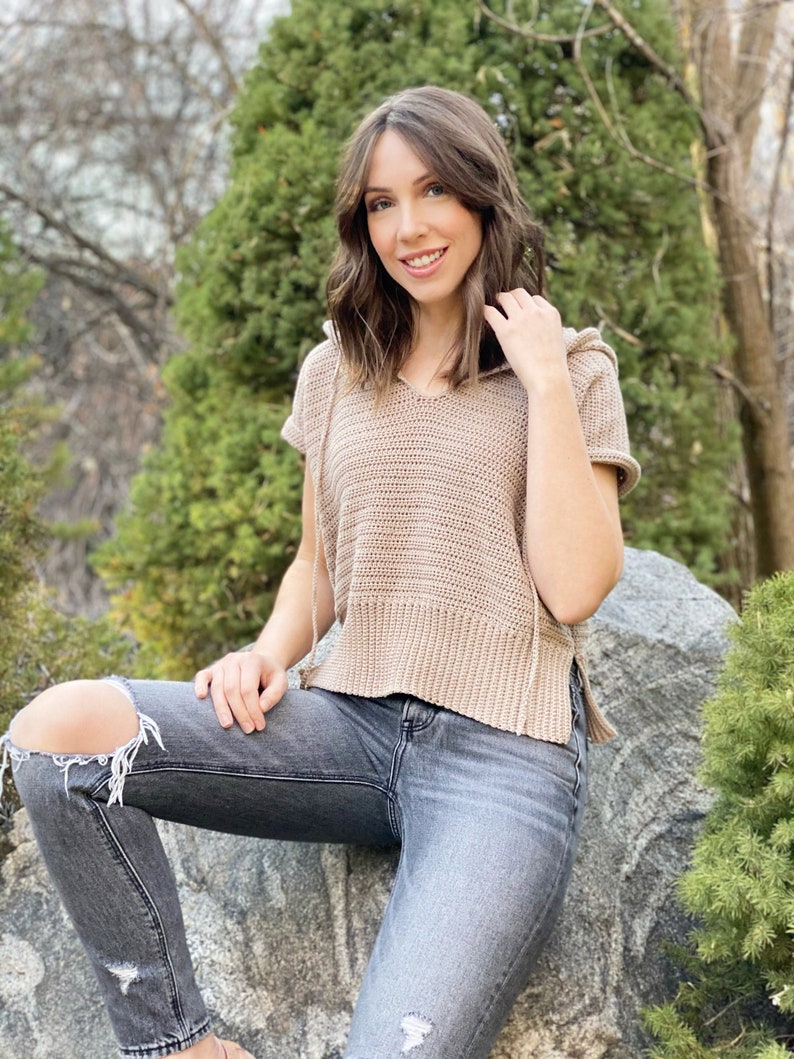 May include: A woman wearing a light brown crocheted hooded top with short sleeves and a pair of dark grey jeans with distressed details.