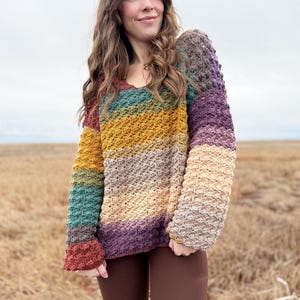 May include: A multi-coloured striped crochet sweater with a v-neck and long sleeves. The sweater is made with a textured stitch and has a relaxed fit.