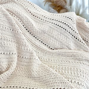 Può includere: Una coperta color crema lavorata all'uncinetto a mano con un design strutturato. La coperta presenta un motivo ripetuto di sezioni piene e traforate, creando una trama visivamente accattivante e accogliente. Il colore neutro la rende adatta a vari stili di arredamento.