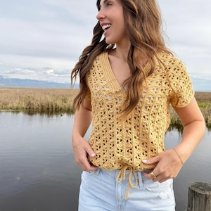 May include: Mustard yellow crochet top with a V-neck and short sleeves. The top features a drawstring waist and a geometric openwork pattern. The model is wearing light blue denim shorts.