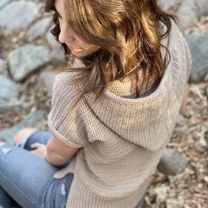 May include: A light brown hooded sweater with short sleeves and a crocheted texture. The sweater is being worn by a person sitting on a bed of rocks and leaves.