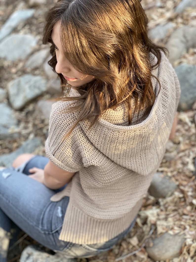 May include: A light brown hooded sweater with short sleeves and a crocheted texture. The sweater is being worn by a person sitting on a bed of rocks and leaves.