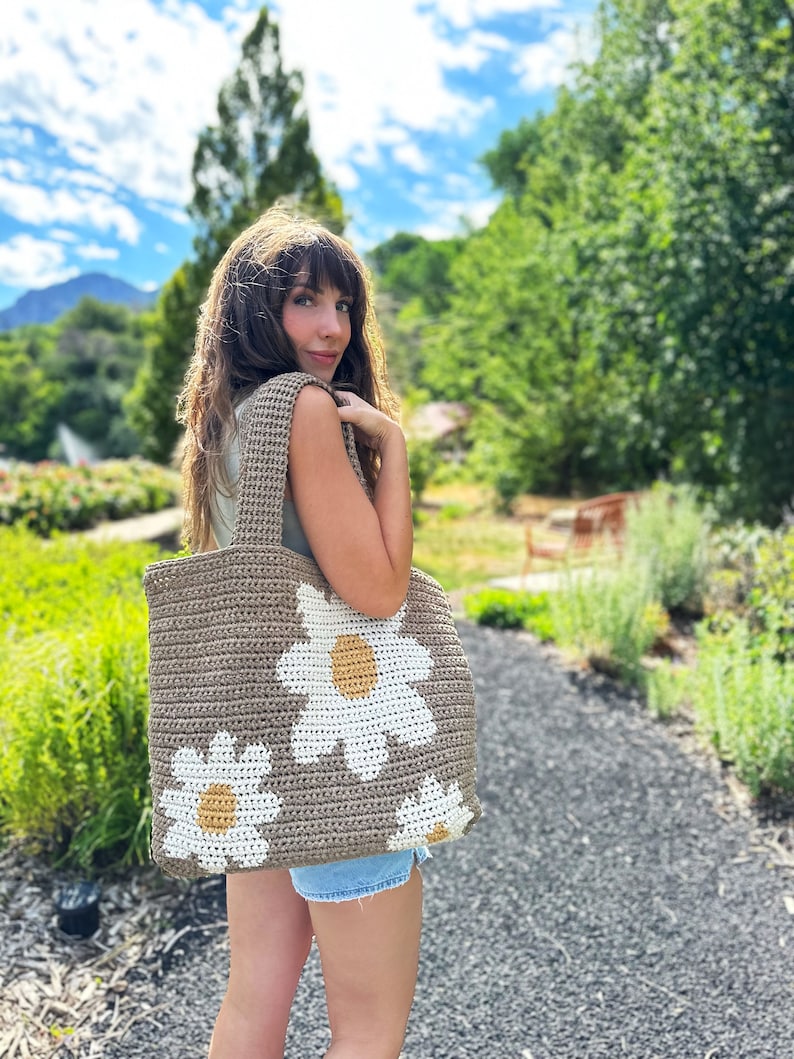May include: A woman is holding a crocheted tote bag with a daisy design. The bag is brown with white and yellow daisies.