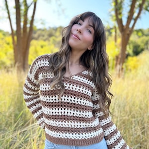 May include: A brown and white striped crochet sweater with long sleeves. The sweater has a V-neck and a loose fit.
