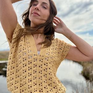 May include: A short-sleeved, mustard yellow crochet top with a V-neck and drawstring waist. The top features an open, geometric pattern. The model is wearing the top outdoors with blue jeans.