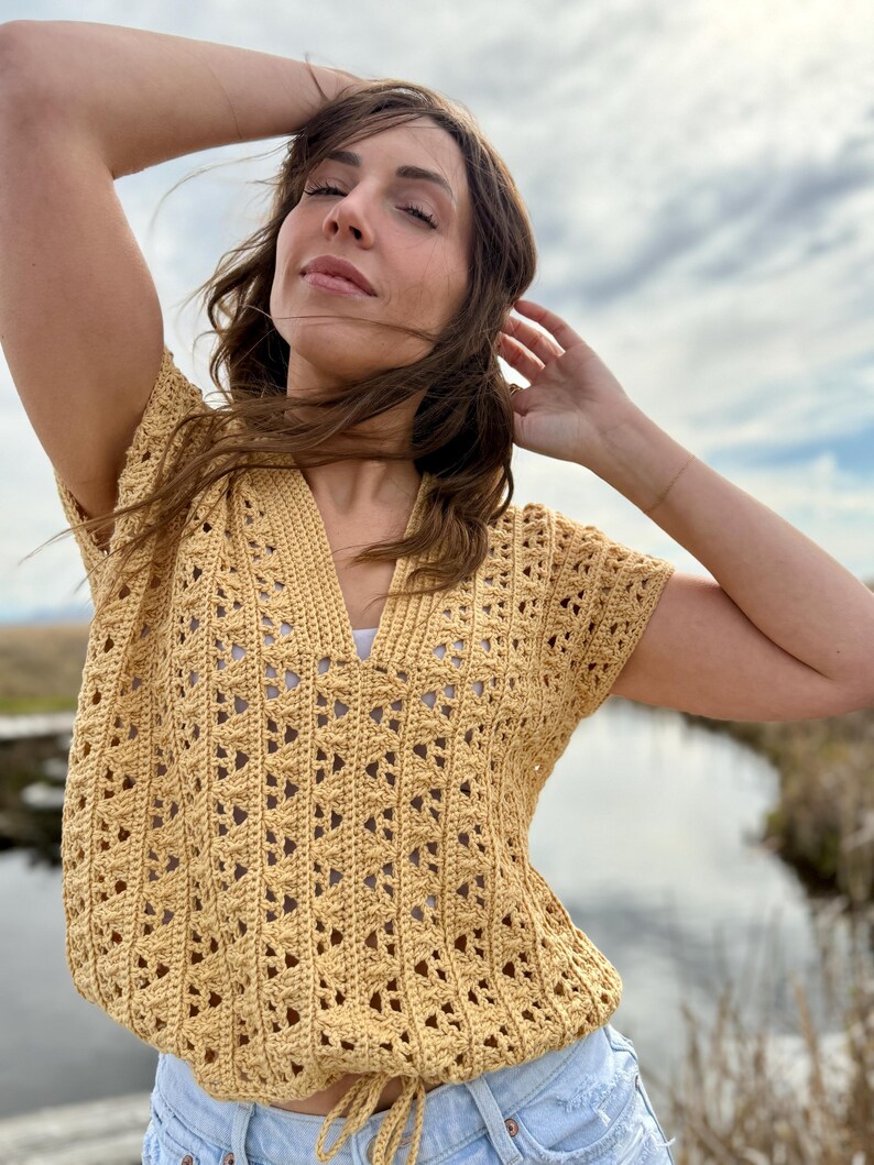 May include: A short-sleeved, mustard yellow crochet top with a V-neck and drawstring waist. The top features an open, geometric pattern. The model is wearing the top outdoors with blue jeans.