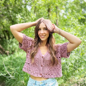 May include: A mauve crochet cardigan with short sleeves and a V-neck. The cropped top features a button-up front and a delicate open weave design. The woman is wearing light blue denim shorts. The background is a lush green forest.