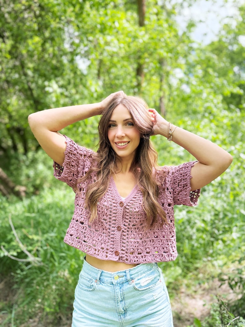 May include: A mauve crochet cardigan with short sleeves and a V-neck. The cropped top features a button-up front and a delicate open weave design. The woman is wearing light blue denim shorts. The background is a lush green forest.