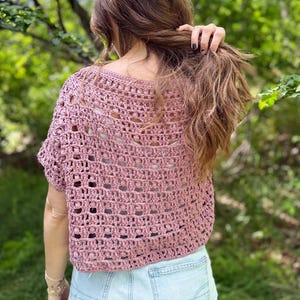 May include: A dusty rose crochet top with a short sleeve design. The top has an open, airy weave pattern. The person is wearing light blue denim shorts. The background is a blurred view of green foliage.