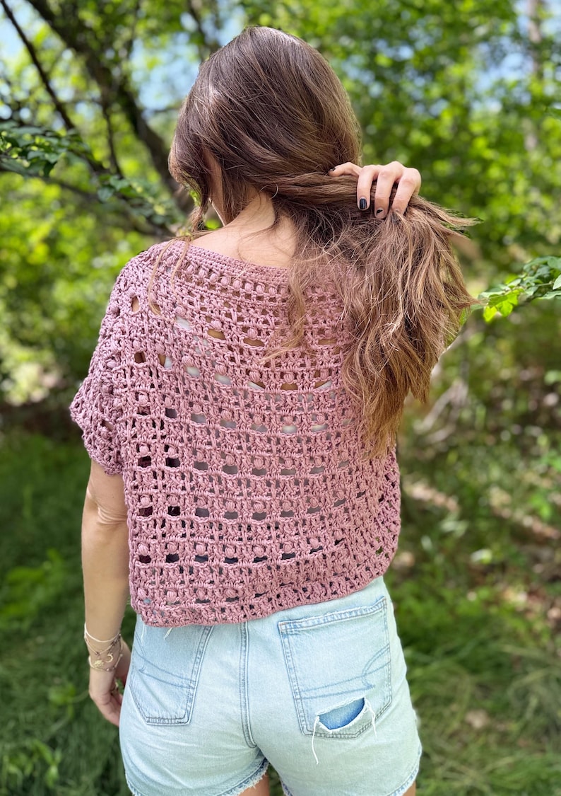 May include: A dusty rose crochet top with a short sleeve design. The top has an open, airy weave pattern. The person is wearing light blue denim shorts. The background is a blurred view of green foliage.