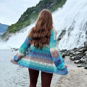May include: A colorful, open-front crochet cardigan in shades of blue, turquoise, and brown. The handmade garment features long sleeves and a striped pattern. The cardigan is worn outdoors near a waterfall.