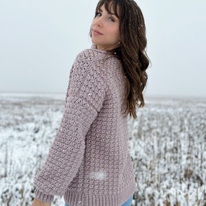 May include: A mauve crocheted sweater with a textured stitch pattern. The sweater has long sleeves and a relaxed fit. The model is wearing the sweater with blue jeans and is standing in a snowy field.
