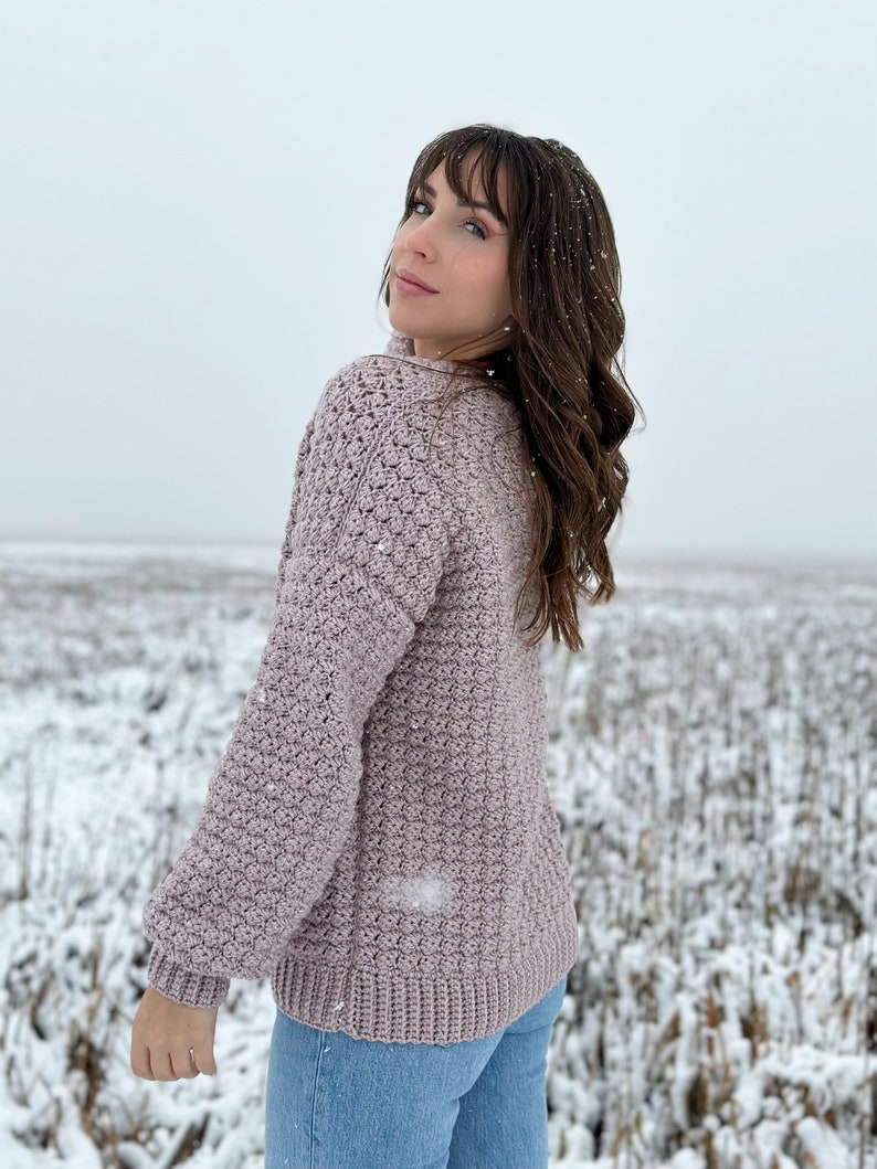 May include: A mauve crocheted sweater with a textured stitch pattern. The sweater has long sleeves and a relaxed fit. The model is wearing the sweater with blue jeans and is standing in a snowy field.
