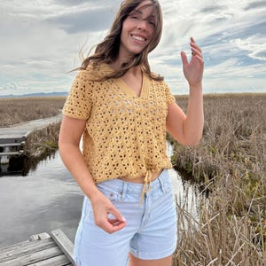 May include: A mustard yellow crochet top with a V-neck and short sleeves. The top has a drawstring at the waist and is paired with light blue denim shorts. The outfit is worn outdoors with a cloudy sky in the background.