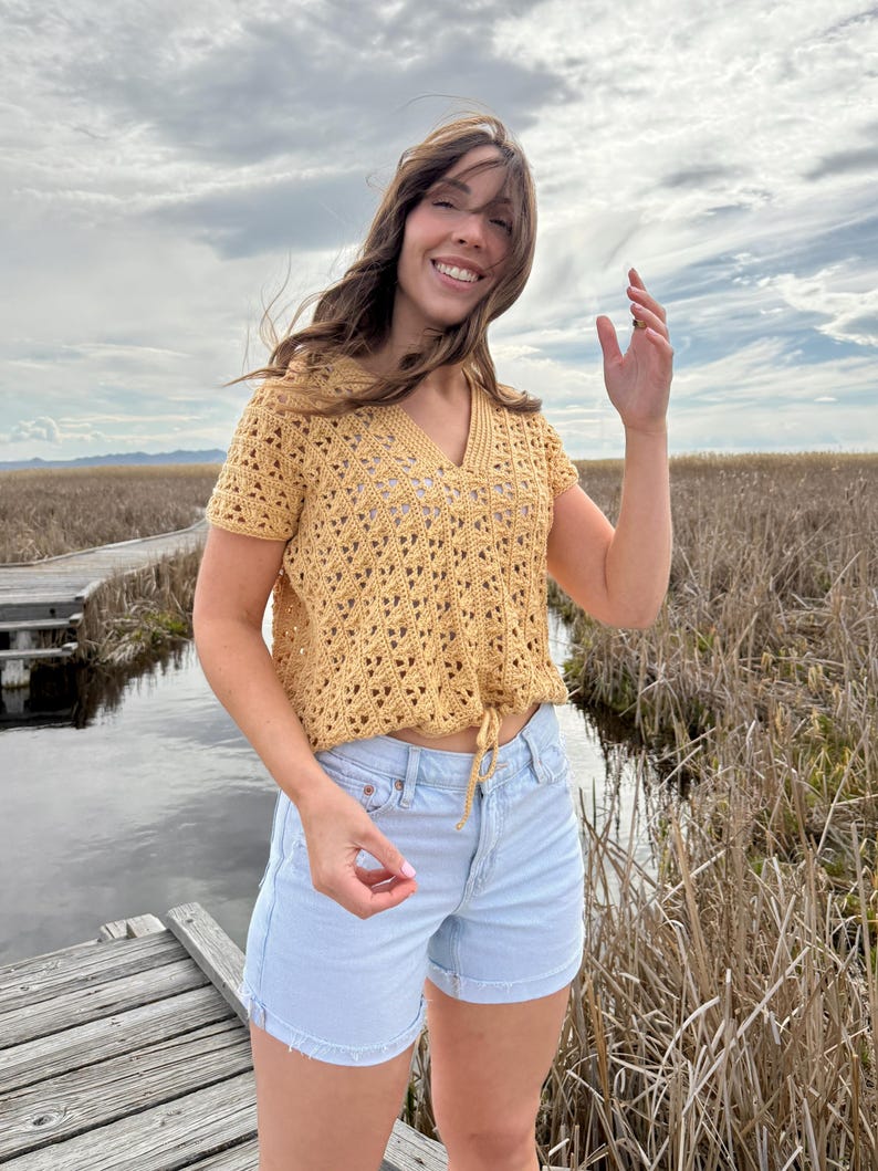 May include: A mustard yellow crochet top with a V-neck and short sleeves. The top has a drawstring at the waist and is paired with light blue denim shorts. The outfit is worn outdoors with a cloudy sky in the background.