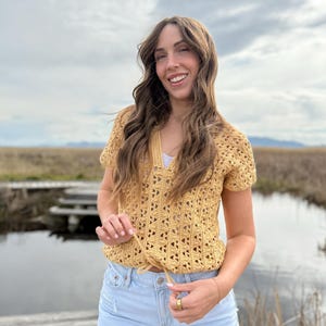May include: Mustard yellow crochet top with a V-neck and short sleeves. The top has a drawstring at the waist and is paired with light blue denim shorts. The outfit is worn outdoors with a natural background.