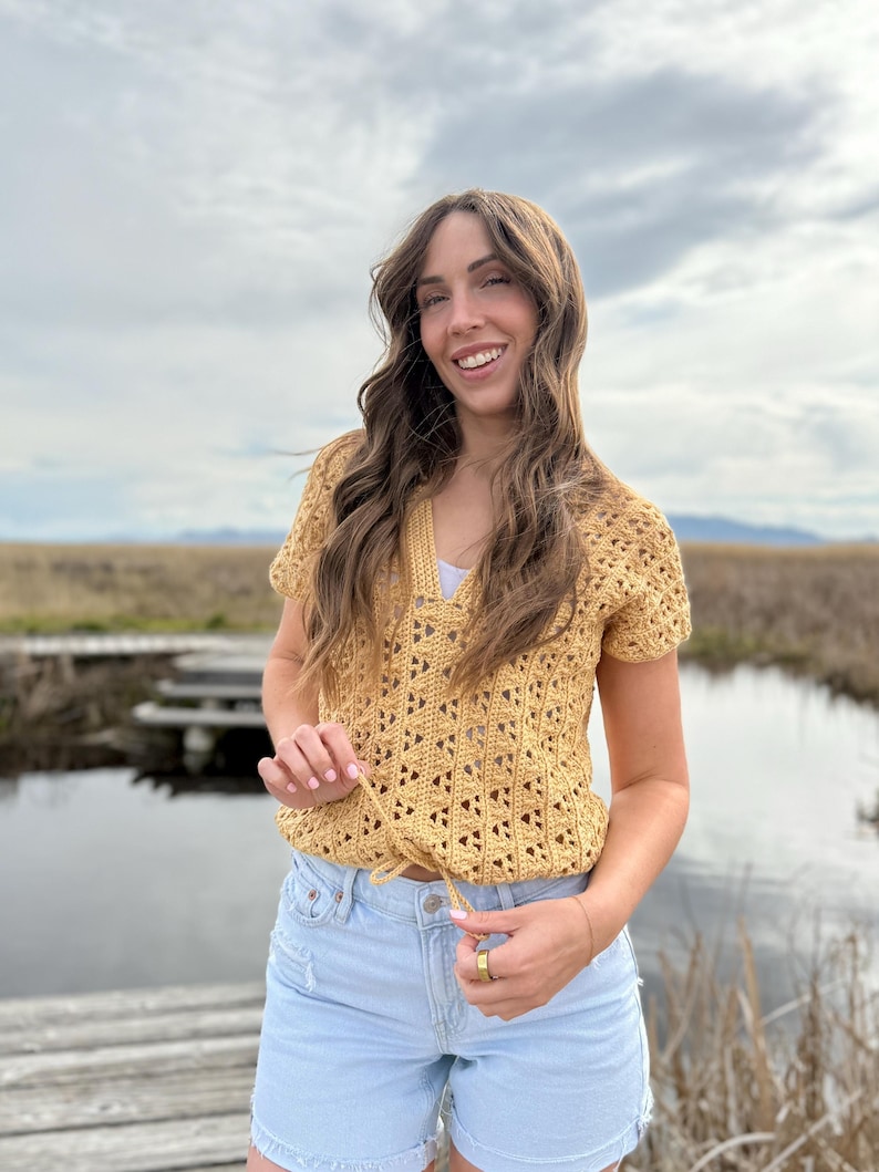 May include: Mustard yellow crochet top with a V-neck and short sleeves. The top has a drawstring at the waist and is paired with light blue denim shorts. The outfit is worn outdoors with a natural background.