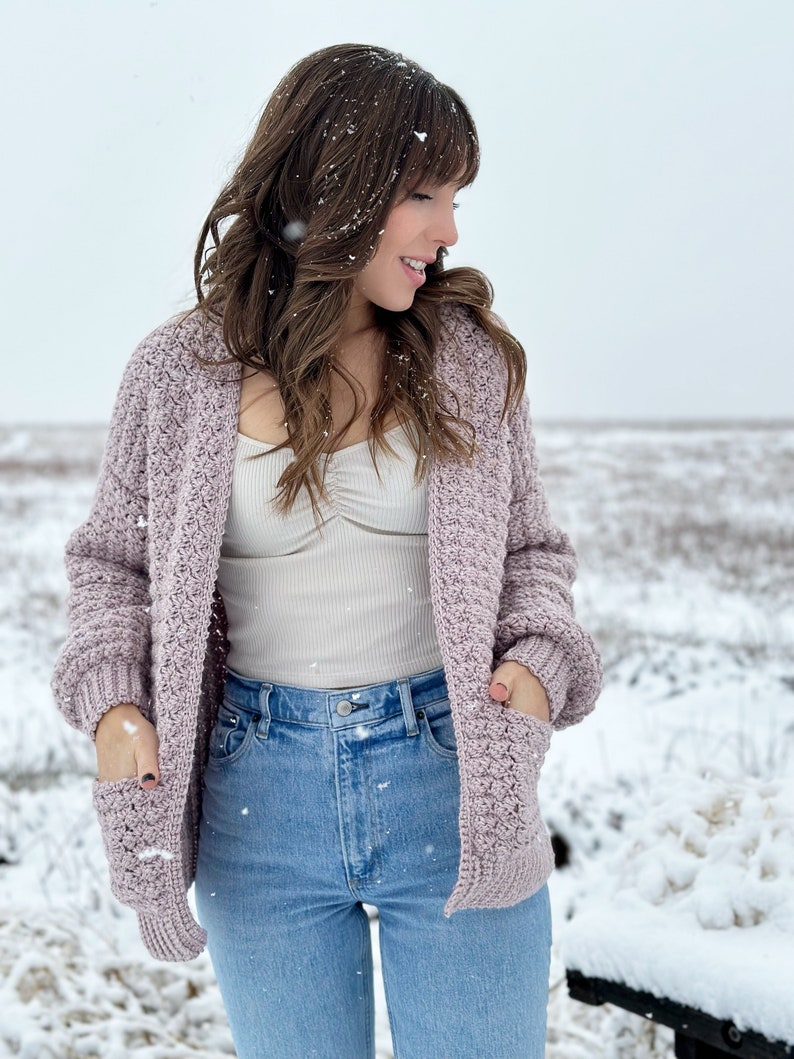 May include: A woman wearing a light purple crocheted cardigan with pockets, a white tank top, and blue jeans. She is standing in a snowy field.