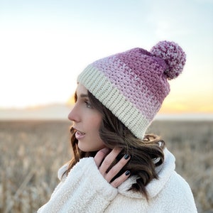 Puede incluir: Una mujer con una chaqueta blanca peluda y un gorro de punto de color rosa y blanco con un pompón blanco.
