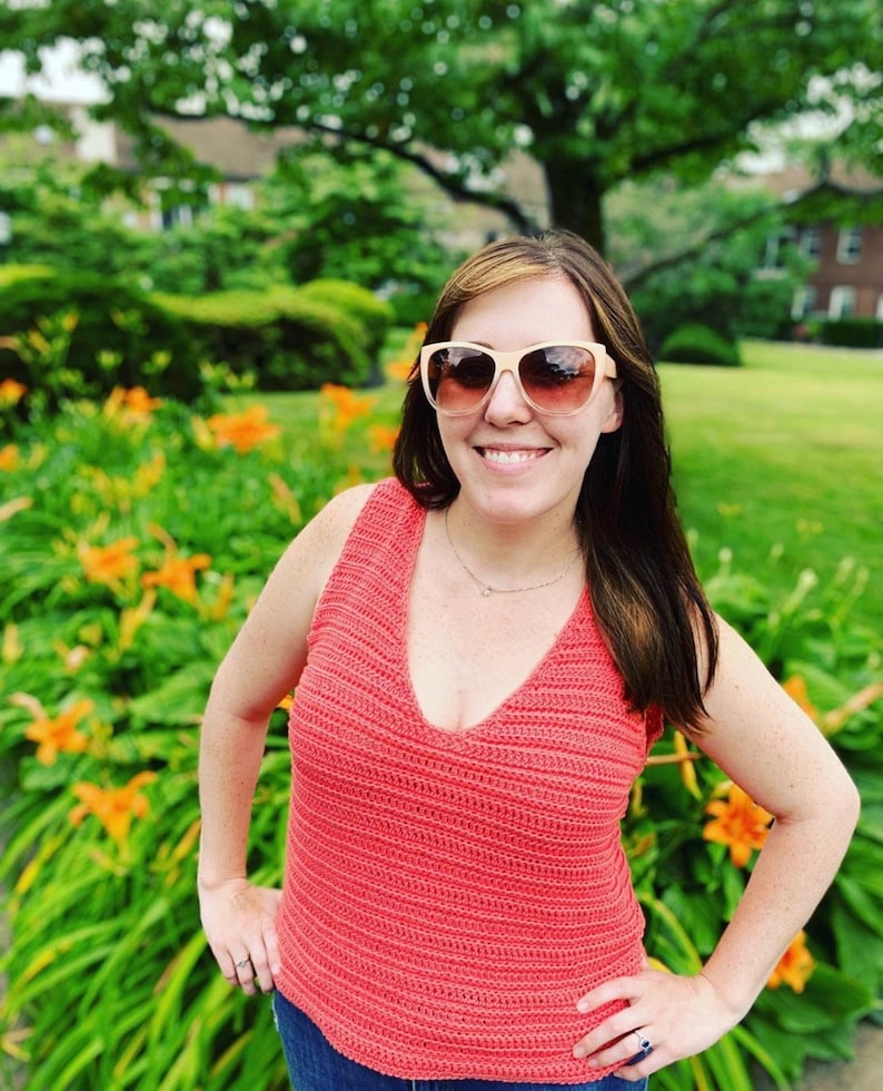 May include: A woman wearing a coral crochet tank top with a deep V-neckline. She is wearing sunglasses and smiling.