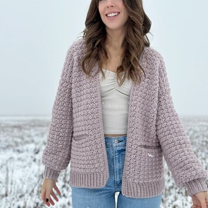 May include: A woman wearing a mauve crocheted cardigan with pockets and blue jeans. She is standing in a snowy field.