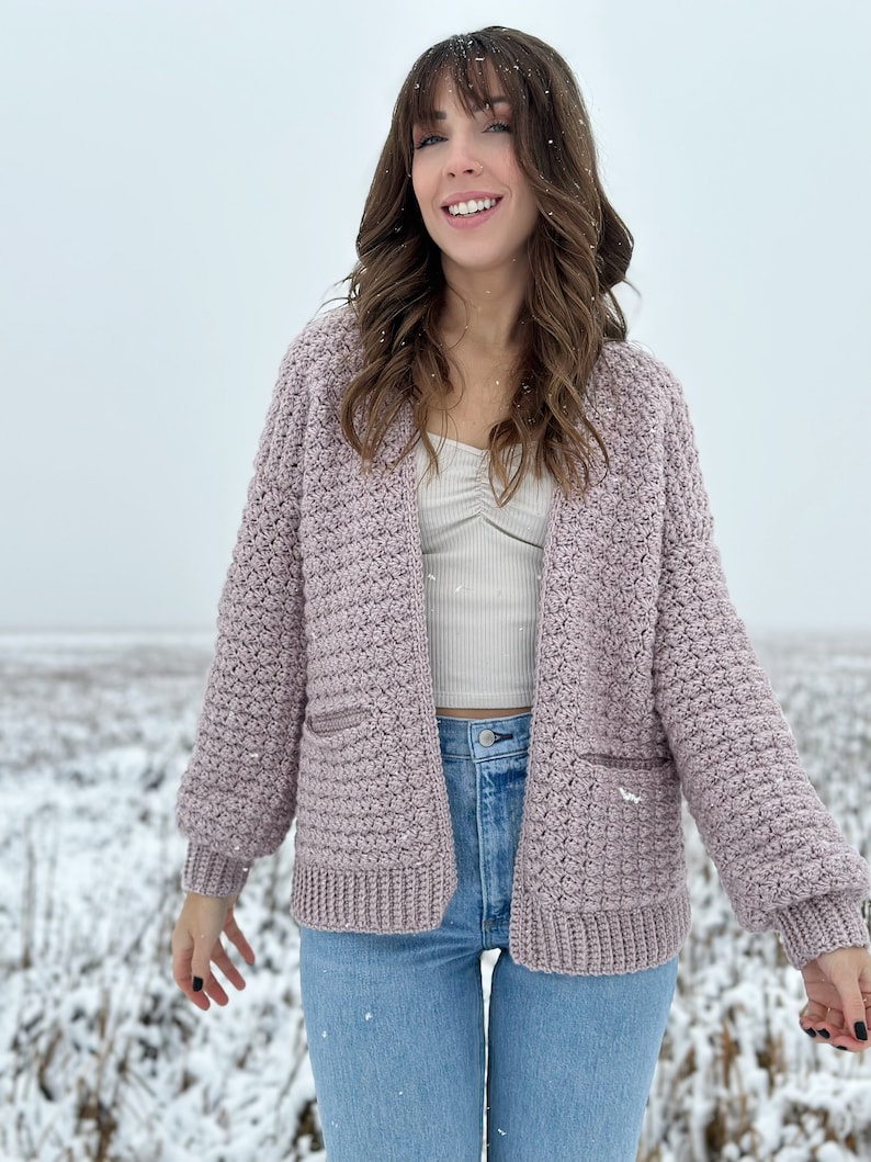 May include: A woman wearing a mauve crocheted cardigan with pockets and blue jeans. She is standing in a snowy field.