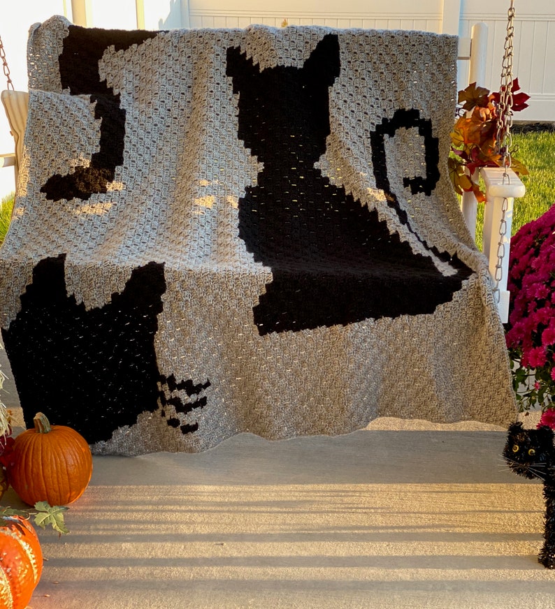 Op de afbeelding: Een grijs gehaakt deken met twee zwarte katten silhouetten. Het deken is gedrapeerd over een witte veranda schommel met pompoenen en herfstbladeren op de achtergrond.