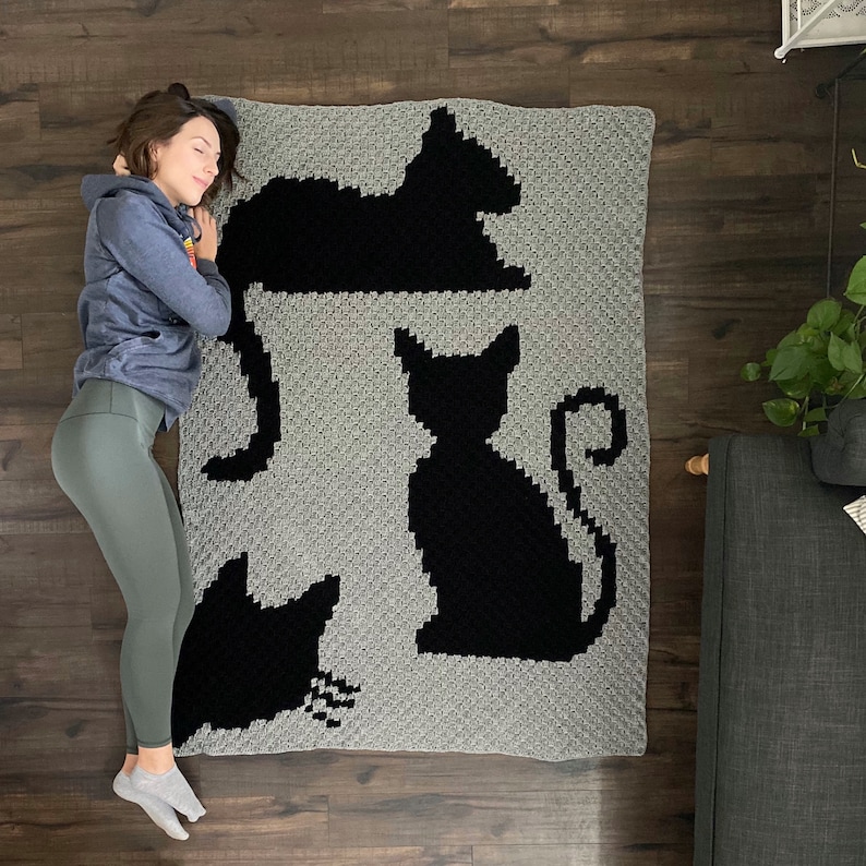 Op de afbeelding: Een grijs gehaakt deken met zwarte katten silhouetten. Het deken heeft drie katten in verschillende poses, waaronder een kat die opgerold ligt, een kat die zit en een kat die staat.