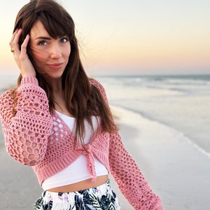 May include: A woman wearing a pink crocheted cardigan, a white tank top, and a floral skirt stands on a sandy beach with the ocean in the background.