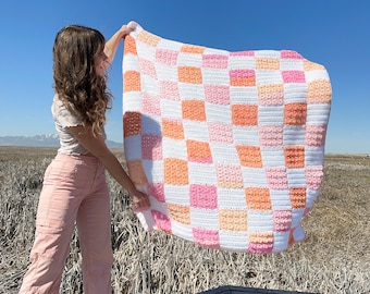 May include: A person holds up a crocheted blanket with a checkered pattern in shades of pink, orange, and white.