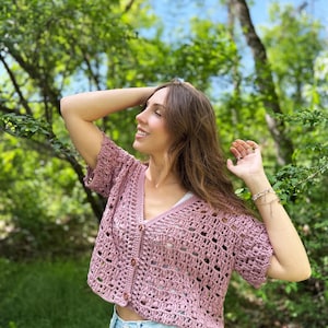 May include: A short-sleeved, dusty rose crochet cardigan with a V-neck and button closure. The open weave design reveals a white top underneath. The cardigan is paired with light blue denim shorts, creating a casual, summery look.