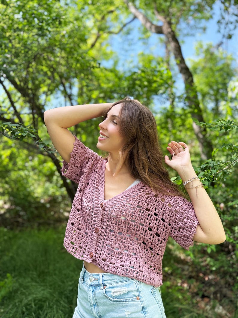 May include: A short-sleeved, dusty rose crochet cardigan with a V-neck and button closure. The open weave design reveals a white top underneath. The cardigan is paired with light blue denim shorts, creating a casual, summery look.